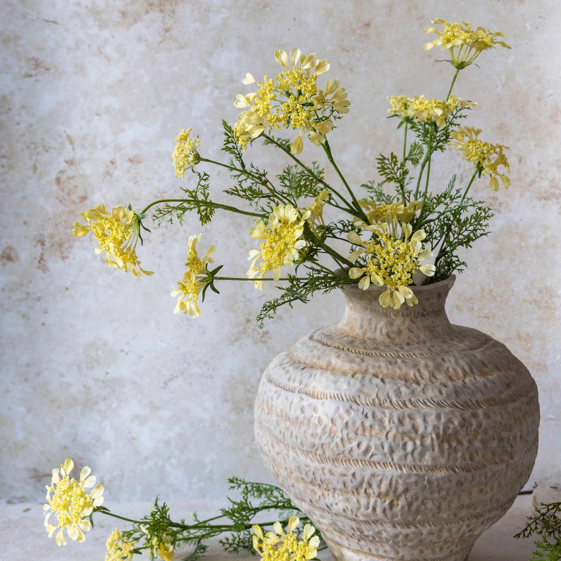 Yellow flowers in a textured stone vase against a light stone wall.